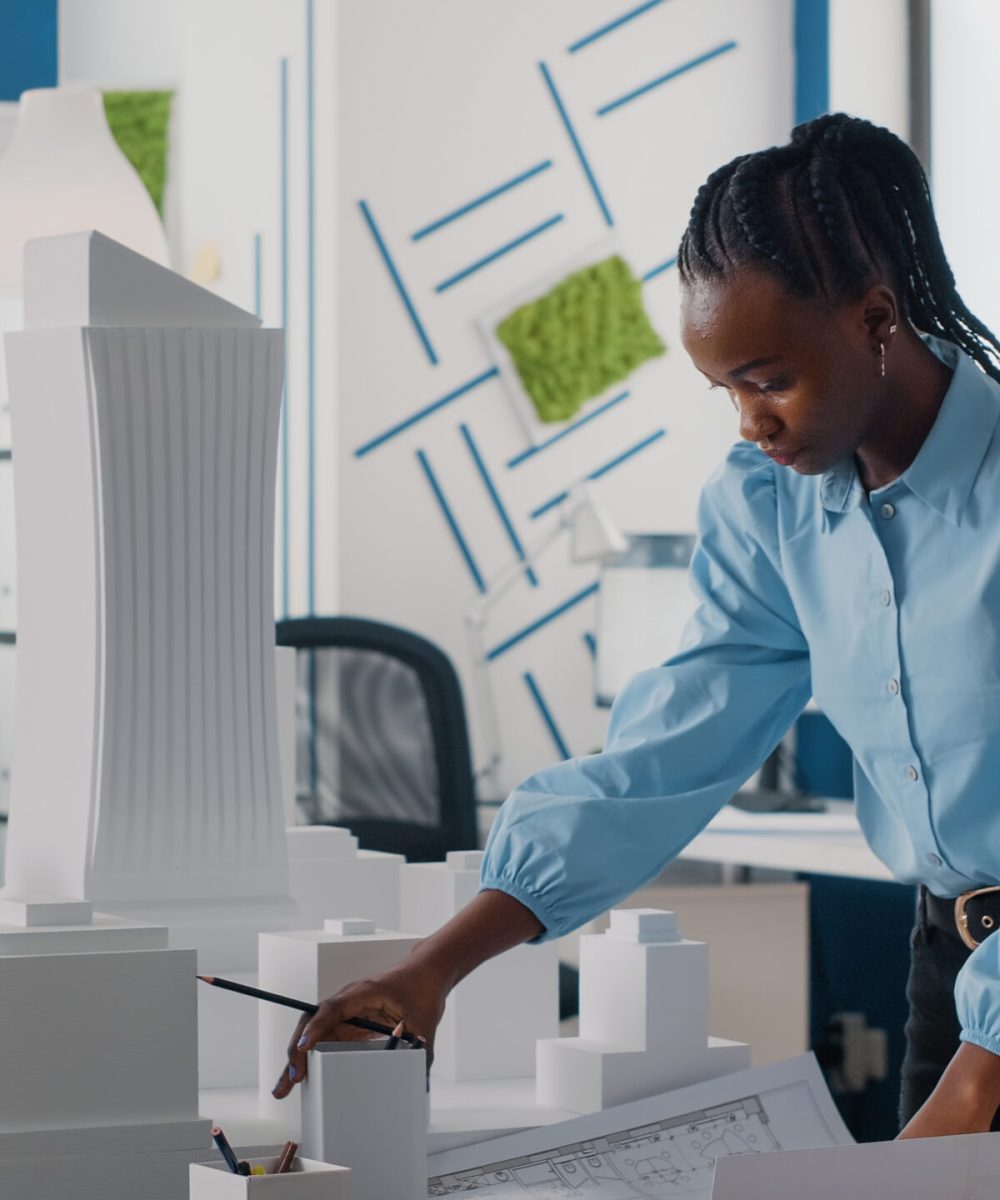African american woman analyzing blueprints plan and building model to work on architecture. Architect working with maquette and layout print to design urban construction for development.